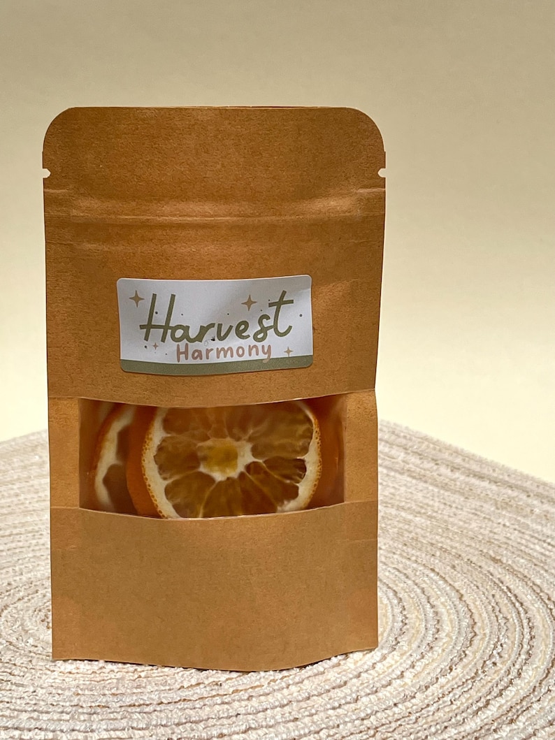 Peut inclure: Un sac en papier brun avec une fen&ecirc;tre transparente montrant des tranches d'orange s&eacute;ch&eacute;es. Le sac porte une &eacute;tiquette blanche avec le texte "Harvest Harmony" en noir.