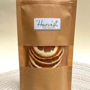 Peut inclure: Un sac en papier brun avec une fen&ecirc;tre transparente montrant des tranches d'orange s&eacute;ch&eacute;es. Le sac porte une &eacute;tiquette blanche avec le texte "Harvest Harmony" en lettres brunes.