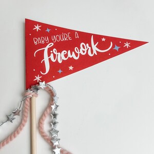 Könnte beinhalten: Ein roter Filz-Wimpel mit dem Text "Baby you're a firework" in weißer Schrift. Der Wimpel ist an einem Holzstab mit einer rosa Garn- und Silberstern-Girlande befestigt.
