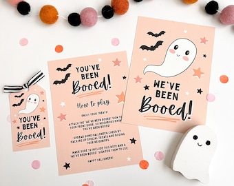 Può includere: Un cartello stampabile di Halloween "You've Been Booed!" con una simpatica illustrazione di un fantasma e istruzioni su come giocare al gioco. Il cartello è rosa con accenti neri e bianchi.