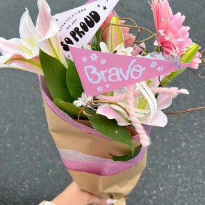 May include: A bouquet of pink and white lilies and pink gerbera daisies wrapped in brown paper with pink tissue paper. Two pink pennants with the text "We are so proud" and "Bravo" are attached to the bouquet.