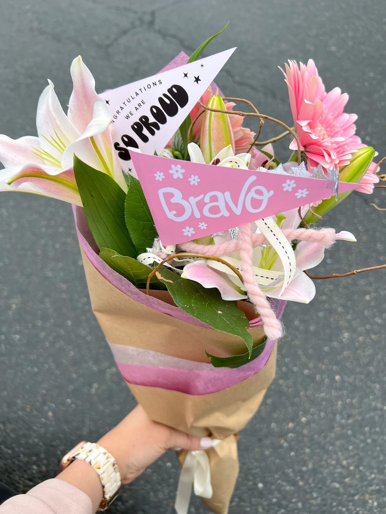 May include: A bouquet of pink and white lilies and pink gerbera daisies wrapped in brown paper with pink tissue paper. Two pink pennants with the text "We are so proud" and "Bravo" are attached to the bouquet.