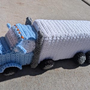 May include: A crocheted toy truck with a blue cab and a white trailer. The truck has black wheels and orange details.