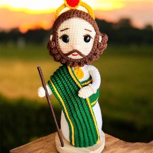May include: A crocheted figure of a saint, featuring a white robe, green and yellow shawl, and a brown beard. The figure holds a wooden staff and wears a gold halo with a red gem. The figure is on a wooden base.
