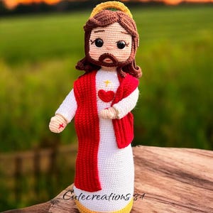 May include: Crocheted doll depicting a religious figure, wearing a white robe, red vest, and a gold halo. The doll has brown hair and a beard, with a red heart and cross on the chest. The doll is handmade.