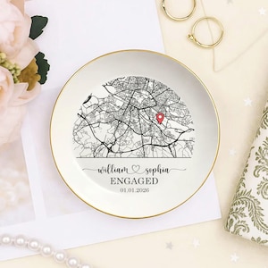 May include: A white ceramic ring dish with a gold rim features a black and white map design. The dish is personalized with the names "William & Sophia", the word "ENGAGED", and the date "01.01.2026". A red pin marks a location on the map.