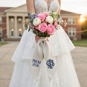 May include: A bouquet of pink roses, white flowers, and greenery tied with a beige ribbon. The ribbon has the text "PROM 2026" and a monogram in navy blue. The bouquet is held by a person wearing a white dress.
