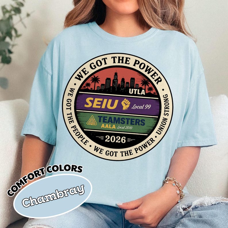 May include: Light blue t-shirt with a circular graphic. The graphic features the text "WE GOT THE POWER" and "WE GOT THE PEOPLE" around a central design. The design includes a cityscape, palm trees, and the text "SEIU", "TEAMSTERS", and "2026". The shirt is labeled "Comfort Colors Chambray".