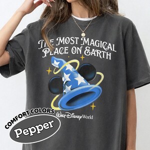 May include: Dark gray t-shirt featuring the text "THE MOST MAGICAL PLACE ON EARTH" above a blue wizard hat with white stars and Mickey Mouse ears. The shirt also has the text "Walt Disney World" and "COMFORT COLORS Pepper".