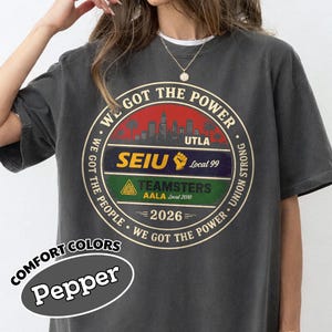 May include: A dark gray t-shirt with a circular graphic that reads "WE GOT THE POWER." The graphic includes the text "UTLA," "SEIU Local 99," and "TEAMSTERS AALA Local 2010." The shirt also features the text "2026" and "COMFORT COLORS Pepper."