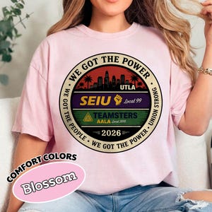 May include: A light pink t-shirt with a circular graphic featuring the text "WE GOT THE POWER" and "UTLA", "SEIU", and "TEAMSTERS" logos. The shirt also has the text "2026" and "COMFORT COLORS Blossom".