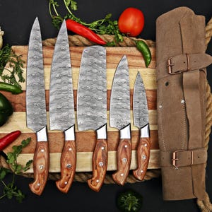 May include: A collection of six kitchen knives with patterned steel blades and wooden handles, presented on a wooden chopping board. A brown leather knife roll is on the right. Fresh vegetables are arranged around the board.