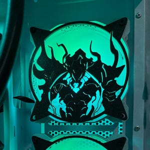 May include: Two computer case fans with black silhouettes of a warrior and a bear, lit by green LED lights. The fans are mounted inside a computer case, creating a vibrant, glowing effect. The overall theme is gaming and computer customization.
