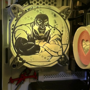 May include: A computer fan with a custom design featuring a muscular figure in a heroic pose. The fan is illuminated with a soft, yellow light, and the design is etched into the fan's surface. The fan is mounted inside a computer case.