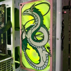 Könnte beinhalten: Ein Computerlüfter mit einem grünen und weißen Drachendesign. Der Lüfter ist in einem weißen Computergehäuse montiert und hat einen rosa Rahmen. Die Lüfterblätter leuchten mit einem hellgelben Licht.