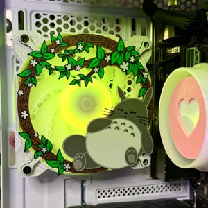 Puede incluir: Un ventilador de computadora con una cubierta decorativa que presenta un personaje de dibujos animados y un diseño floral. Las aspas del ventilador brillan con una luz amarilla. La cubierta incluye una rama marrón con hojas verdes y flores blancas. Un segundo componente tiene un corazón rosa.