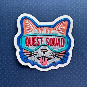 May include: Embroidered iron-on patch featuring a cartoon cat wearing blue and turquoise ski goggles with the text "QUEST SQUAD" in red.