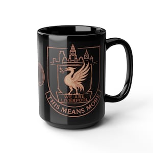 Peut inclure: Mug en céramique noire avec un emblème de l'oiseau Liver de couleur cuivre, le texte "WE ARE LIVERPOOL" et "THIS MEANS MORE", et une silhouette de ville. La tasse a une grande anse.
