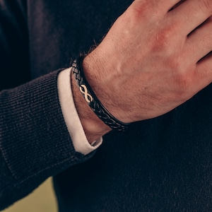 Könnte beinhalten: Ein schwarzes Lederarmband mit einem goldenen Unendlichkeits-Anhänger.