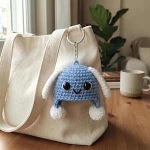 May include: A blue crocheted hat keychain with white ear flaps and two white pom-poms. The keychain is attached to a silver ring and hangs in front of a beige tote bag. The hat has a smiling face with black eyes.