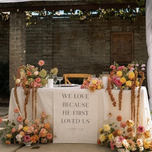 May include: A rectangular table covered with a white linen cloth featuring a vertical runner printed with the text WE LOVE BECAUSE HE FIRST LOVED US 1 JOHN 4:19. The table is decorated with large floral arrangements in shades of pink, yellow, and orange.