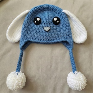 May include: A handmade blue crochet hat with white bunny ears, black eyes, and a smiling mouth. The hat has braided ties with white pom-poms. This whimsical hat is perfect for children and adds a playful touch to any outfit.