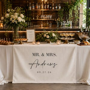 May include: A cream-colored fabric table runner featuring the black printed text MR. & MRS. Andrews and the date 09.27.26. The runner is draped over a white tablecloth on a table decorated with floral arrangements, glassware, and appetizers.