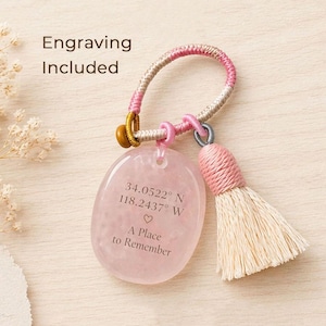 May include: A pink gemstone keyring with a tassel. The keyring has a loop with pink and beige accents, a gold-coloured bead, and a pink gemstone pendant engraved with coordinates and the phrase "A Place to Remember". The text "Engraving Included" is also visible.