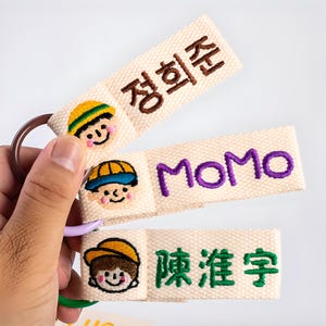 May include: Three beige fabric keychains with embroidered text and cartoon faces. The top keychain has brown text, the middle keychain has purple text, and the bottom keychain has green text. Each keychain has a metal ring.