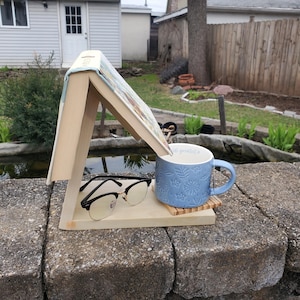 Può includere: Un porta libri in legno con un libro appoggiato, una tazza in ceramica blu con un motivo floreale e un paio di occhiali neri su uno scaffale in legno.