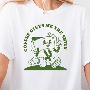Può includere: T-shirt bianco con grafica verde di una tazza di caffè dei cartoni animati. La tazza di caffè fa il pollice in su e il testo recita "COFFEE GIVES ME THE SHITS". Il disegno è centrato sulla parte anteriore della maglietta.