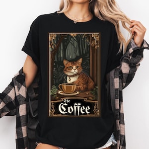 May include: Black t-shirt with a detailed illustration of a cat and a coffee cup. The cat is brown and orange, wearing a collar. The image includes the text "The Coffee" in a decorative font, set against a forest backdrop.