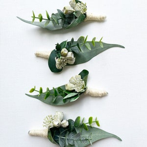 Può includere: Quattro piccoli bouquet di verde e fiori bianchi legati con spago. Ogni bouquet presenta una grande foglia verde.