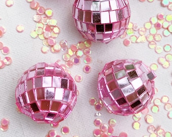 May include: Several small, pink disco ball ornaments with mirrored tiles are scattered across a white surface. Iridescent sequins in shades of pink and yellow surround the disco balls, creating a festive and decorative scene.