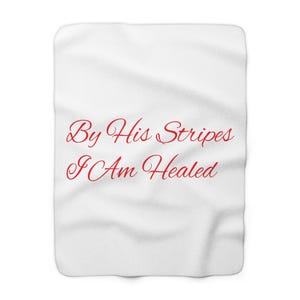 May include: A white blanket featuring the red text "By His Stripes I Am Healed" in a script font. The blanket has a soft, textured appearance.