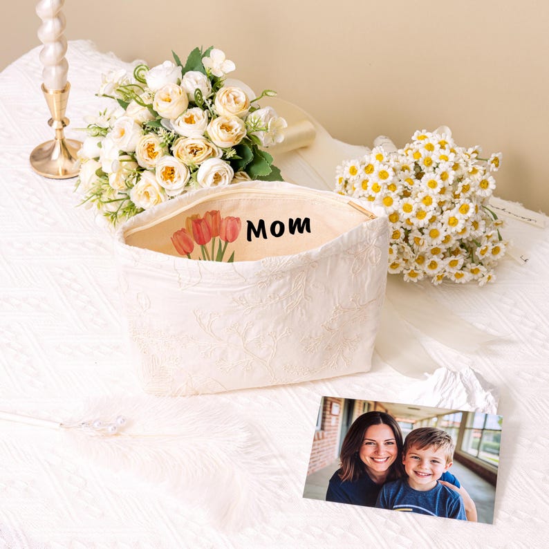 K&ouml;nnte beinhalten: Eine wei&szlig;e gesteppte Rei&szlig;verschlusstasche mit einem Blumendesign und dem Wort "MOM" auf der Vorderseite. Die Tasche liegt auf einer wei&szlig;en Oberfl&auml;che mit einem Strau&szlig; wei&szlig;er Blumen und einem Foto einer Frau und eines Kindes.
