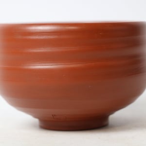 May include: A terracotta ceramic bowl with a rounded shape and a smooth, slightly textured surface. The bowl has a deep red color and is handmade.