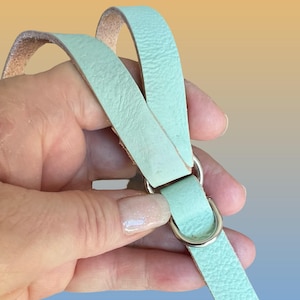 Op de afbeelding: Een close-up van een mintgroene leren riem met een zilveren metalen D-ring en een zilveren metalen ring. De riem wordt door een hand vastgehouden tegen een gradi&euml;nt achtergrond van geel, oranje en blauw.