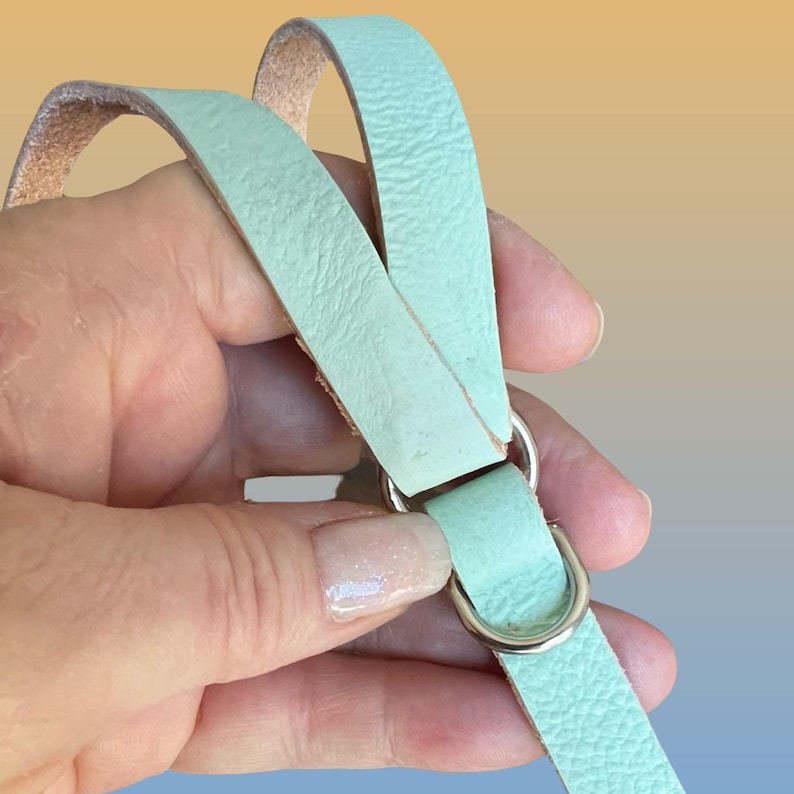 Op de afbeelding: Een close-up van een mintgroene leren riem met een zilveren metalen D-ring en een zilveren metalen ring. De riem wordt door een hand vastgehouden tegen een gradi&euml;nt achtergrond van geel, oranje en blauw.