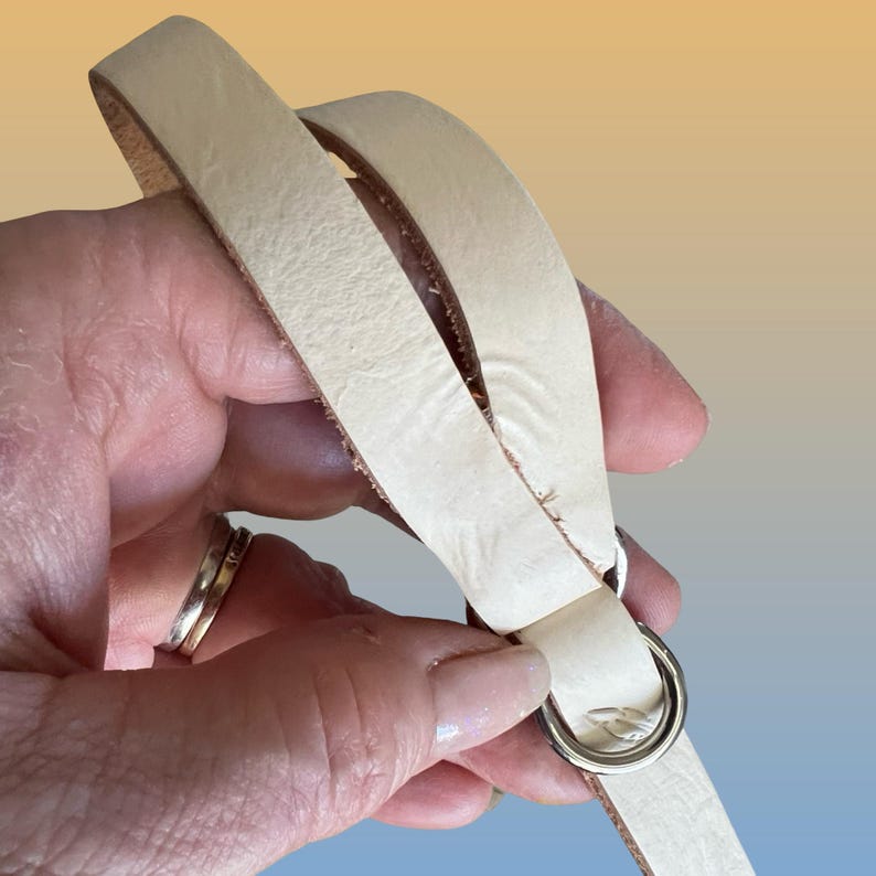 Op de afbeelding: Een cr&egrave;mekleurige leren riem met een zilverkleurige metalen ring. De riem wordt in een hand gehouden, wat de textuur en details van het leer laat zien. De achtergrond heeft een verloop van geel en blauw.