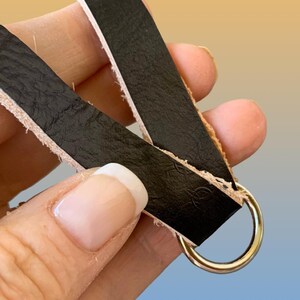 Op de afbeelding: Close-up van zwarte leren bandjes met een goudkleurige metalen ring. Het leer heeft een getextureerd oppervlak en wordt op zijn plaats gehouden door een goudkleurige metalen ring. De randen van het leer zijn onafgewerkt.