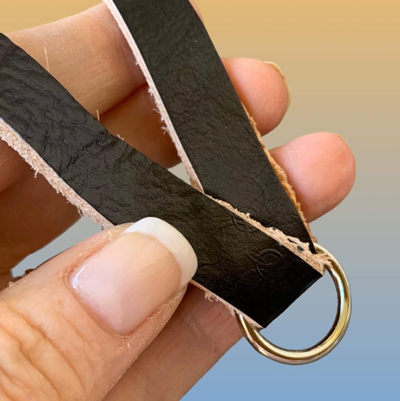 Op de afbeelding: Close-up van zwarte leren bandjes met een goudkleurige metalen ring. Het leer heeft een getextureerd oppervlak en wordt op zijn plaats gehouden door een goudkleurige metalen ring. De randen van het leer zijn onafgewerkt.