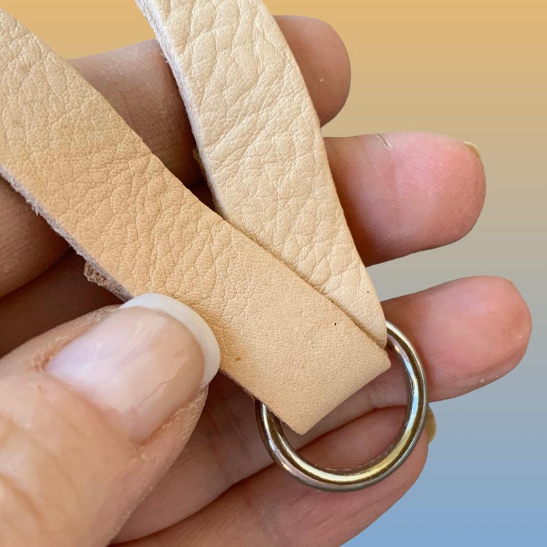 Op de afbeelding: Close-up van een lichtbruine leren riem met een zilveren metalen ring. Het leer heeft een getextureerd oppervlak en wordt in een hand gehouden tegen een gradi&euml;ntachtergrond van geel, oranje en blauw.