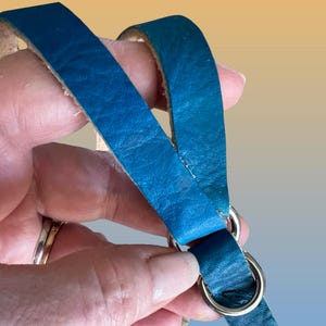Op de afbeelding: Close-up van teal leren riemen verbonden door zilveren metalen ringen. Het leer heeft een getextureerd uiterlijk en de ringen zijn cirkelvormig. De achtergrond heeft een verloop van geel en oranje.