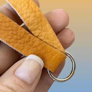 Op de afbeelding: Close-up van een leren sleutelhanger met een zilveren ring. De sleutelhanger heeft twee stroken getextureerd, goudgeel leer. De achtergrond heeft een verloop van geel, blauw en wit.