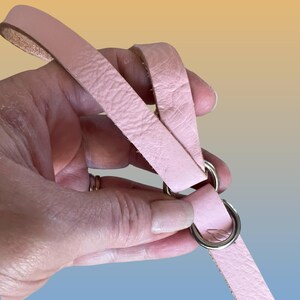 Op de afbeelding: Een lichtroze leren riem met twee zilveren metalen ringen. De riem wordt in een hand gehouden, waardoor de textuur van het leer zichtbaar is. De achtergrond heeft een verloop van geel, oranje en blauw.