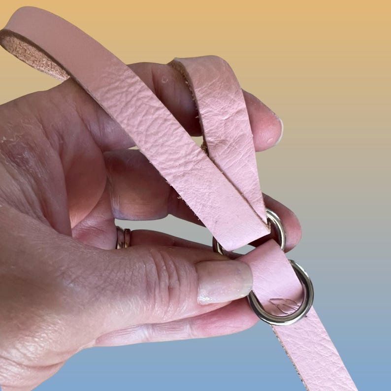Op de afbeelding: Een lichtroze leren riem met twee zilveren metalen ringen. De riem wordt in een hand gehouden, waardoor de textuur van het leer zichtbaar is. De achtergrond heeft een verloop van geel, oranje en blauw.