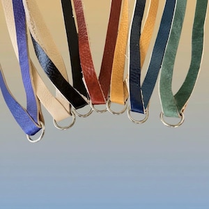 Op de afbeelding: Een verzameling leren riemen in verschillende kleuren, waaronder blauw, cr&egrave;me, zwart, rood en groen, elk bevestigd aan een zilveren metalen ring. De riemen zijn geplaatst tegen een gradi&euml;nt achtergrond.
