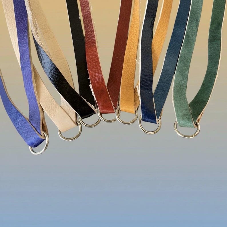 Op de afbeelding: Een verzameling leren riemen in verschillende kleuren, waaronder blauw, cr&egrave;me, zwart, rood en groen, elk bevestigd aan een zilveren metalen ring. De riemen zijn geplaatst tegen een gradi&euml;nt achtergrond.