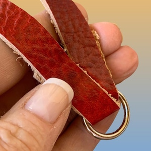 Op de afbeelding: Close-up van een rode leren riem met een goudkleurige metalen ring. Het leer heeft een textuuroppervlak en wordt op zijn plaats gehouden door een metalen ring.
